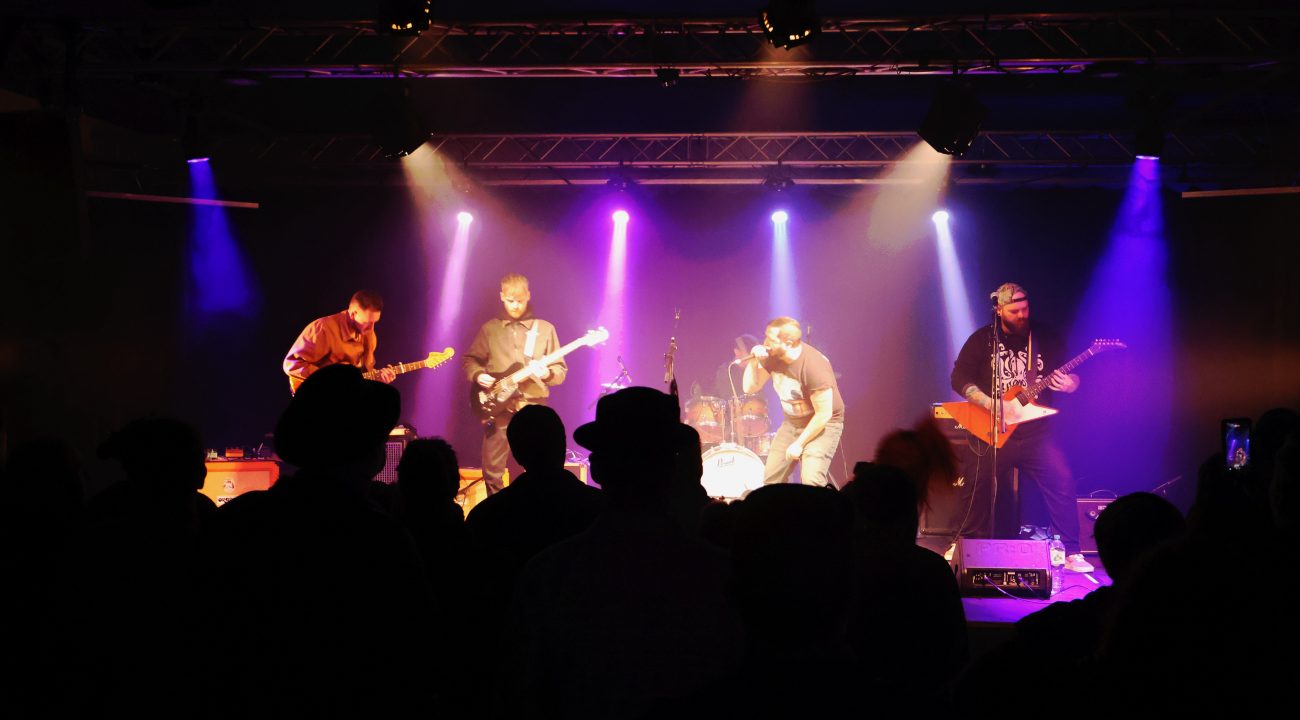 Band performing on stage with colorful lights, silhouetted audience in the foreground.