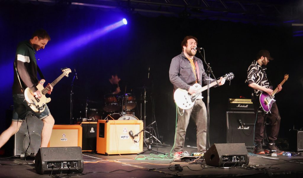 A band performing on stage with colorful lights and various instruments, including guitars, drums, and amplifiers.