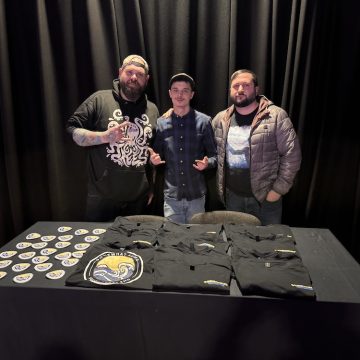 Three people standing behind a table with shirts and stickers, in front of a dark curtain.