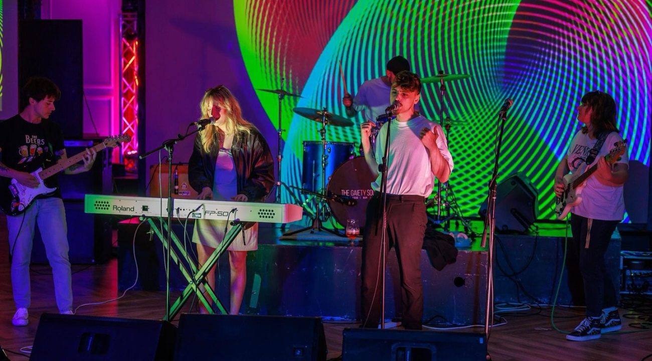 A band performs energetically on stage with colorful, psychedelic light effects in the background.