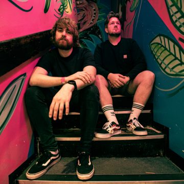 Two men sit on colorful stairs with leafy wall art; one leans forward, the other sits relaxed, both wearing casual clothes.