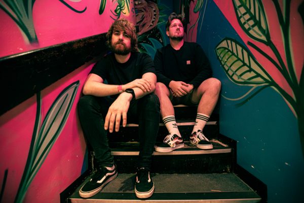 Two men sit on colorful stairs with leafy wall art; one leans forward, the other sits relaxed, both wearing casual clothes.